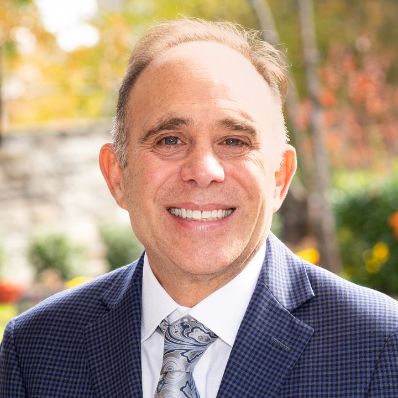 Larry Finkelstein smiling in a professional headshot wearing a navy checked blazer and paisley tie outdoors with autumn foliage