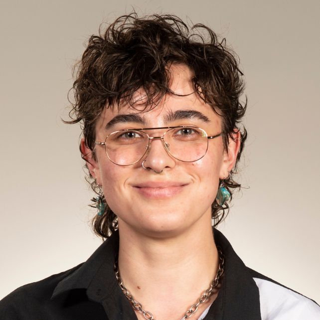 Lee Simmons smiling in a headshot wearing glasses, turquoise earrings, and a chain necklace against a neutral background
