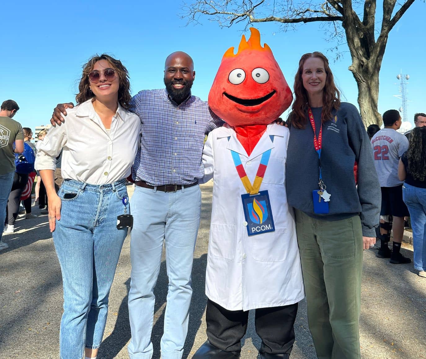 Two staff members pose with the PCOM flame mascot in a white coat at a sunny outdoor campus event