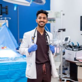 Medical student in a white coat, scrubs, and blue gloves smiles in a clinical simulation lab