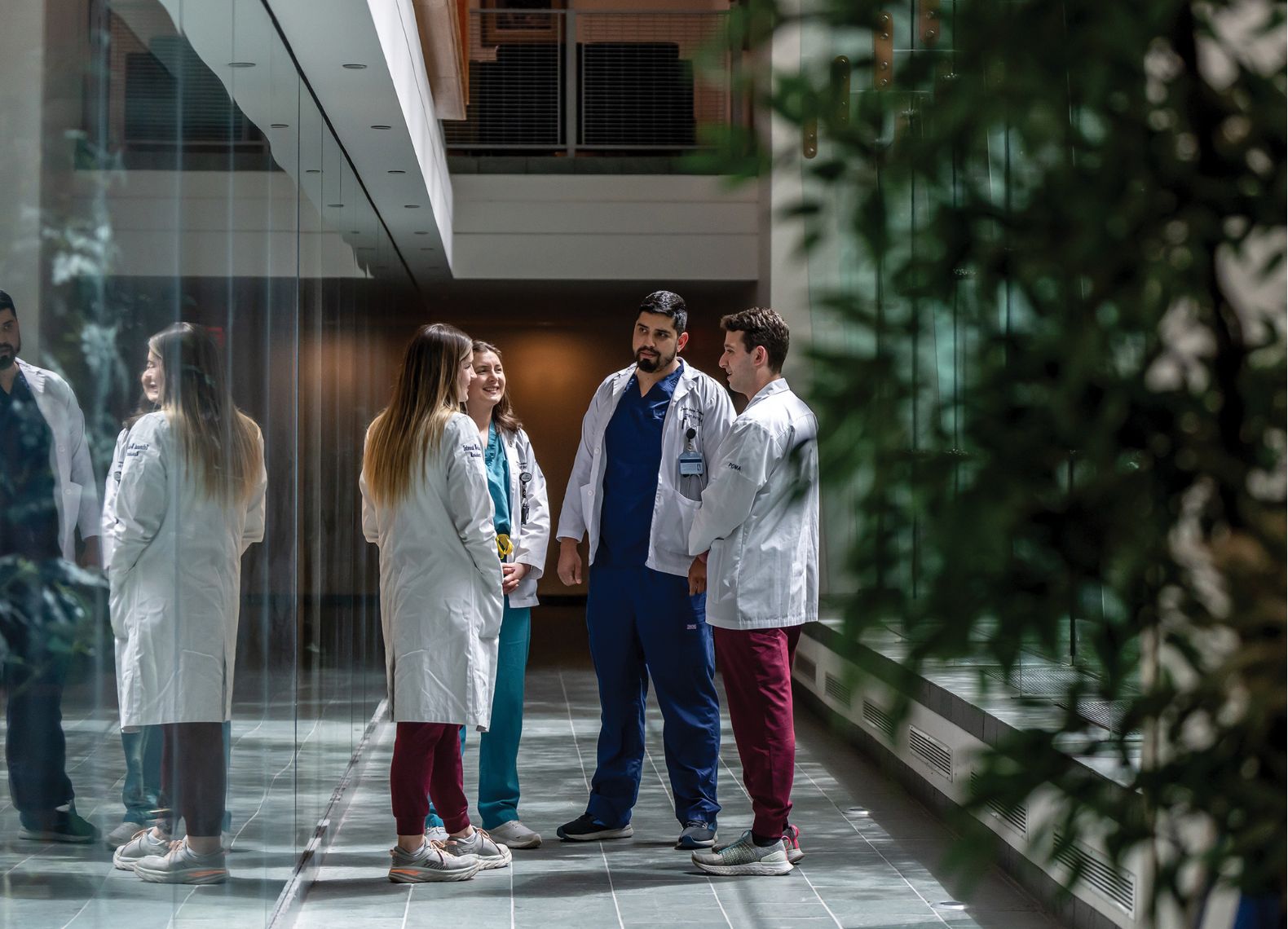 Three medical students in white coats and scrubs talk together in a modern glass-walled campus hallway