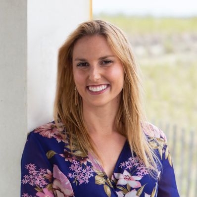 Melanie Garthwaite smiling in a headshot wearing a blue floral top with a coastal backdrop
