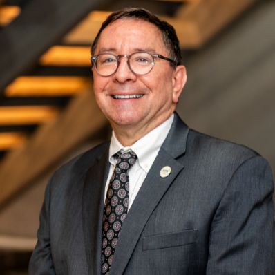 Michael Becker smiling in a professional headshot wearing a gray suit, patterned tie, and glasses in a modern building lobby