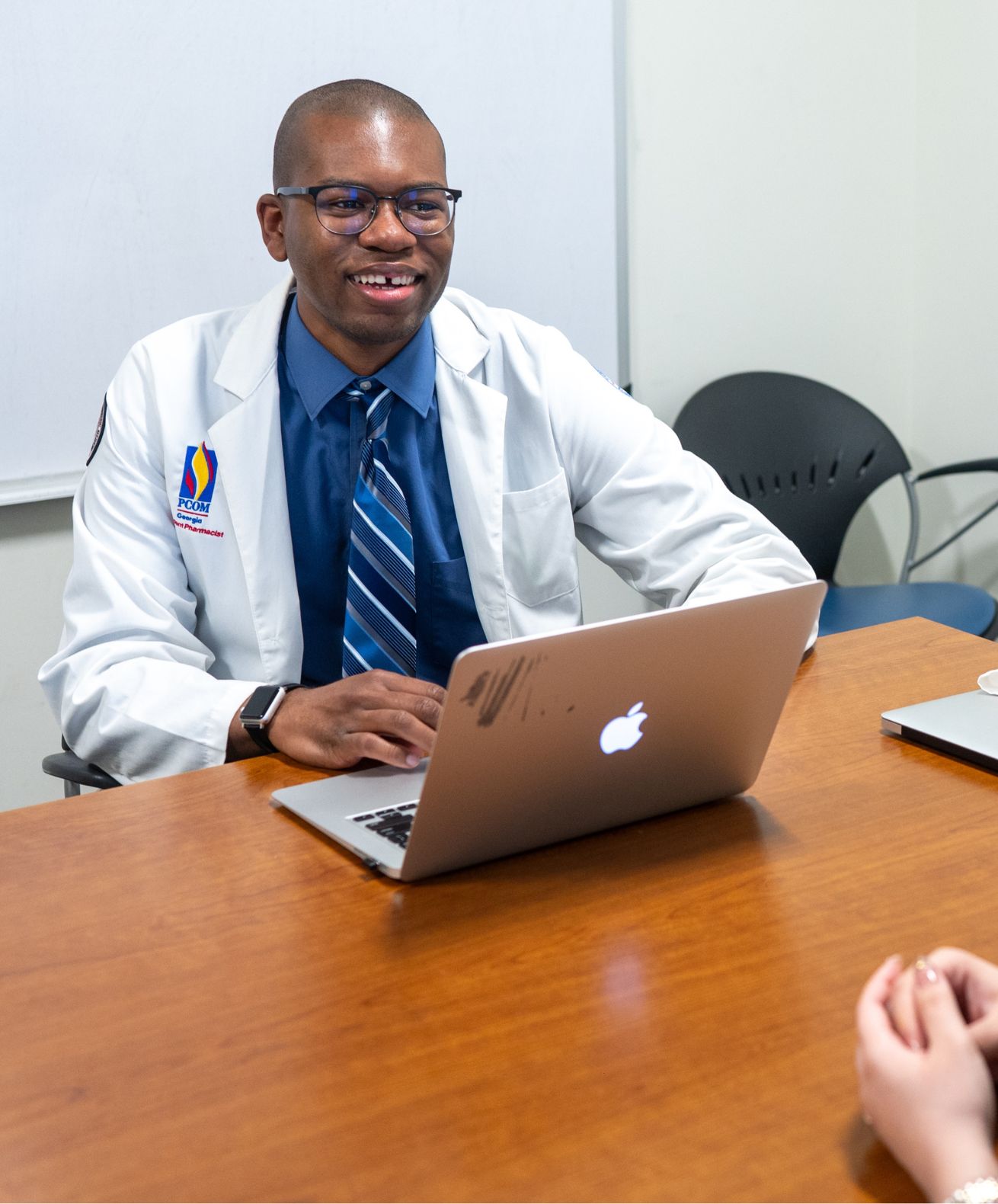 PCOM pharmacy student in a white coat smiles while working on a laptop at a conference table