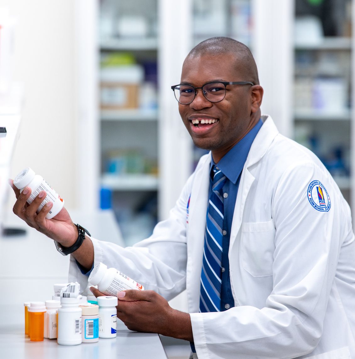Smiling pharmacy student in a PCOM white coat holds medication bottles at a pharmacy lab counter