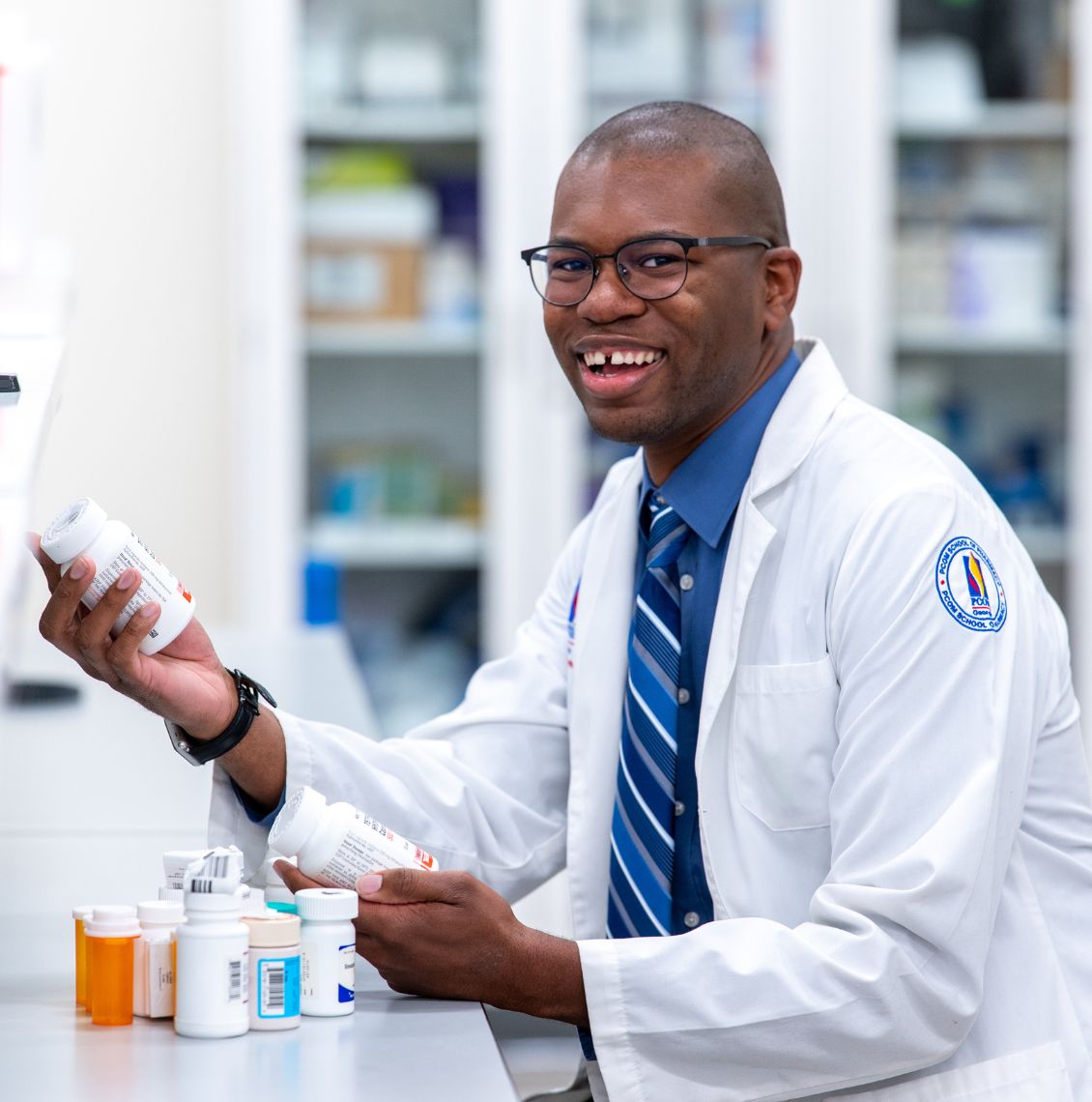 Smiling pharmacy student in a PCOM white coat holds medication bottles at a pharmacy lab counter