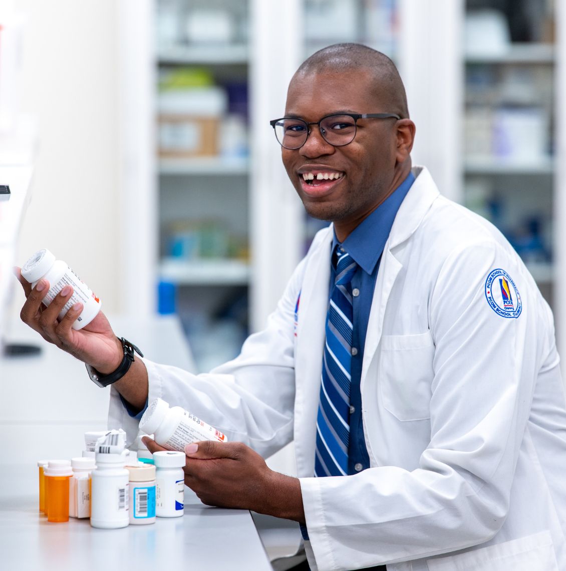 Smiling pharmacy student in a PCOM white coat holds medication bottles at a pharmacy lab counter