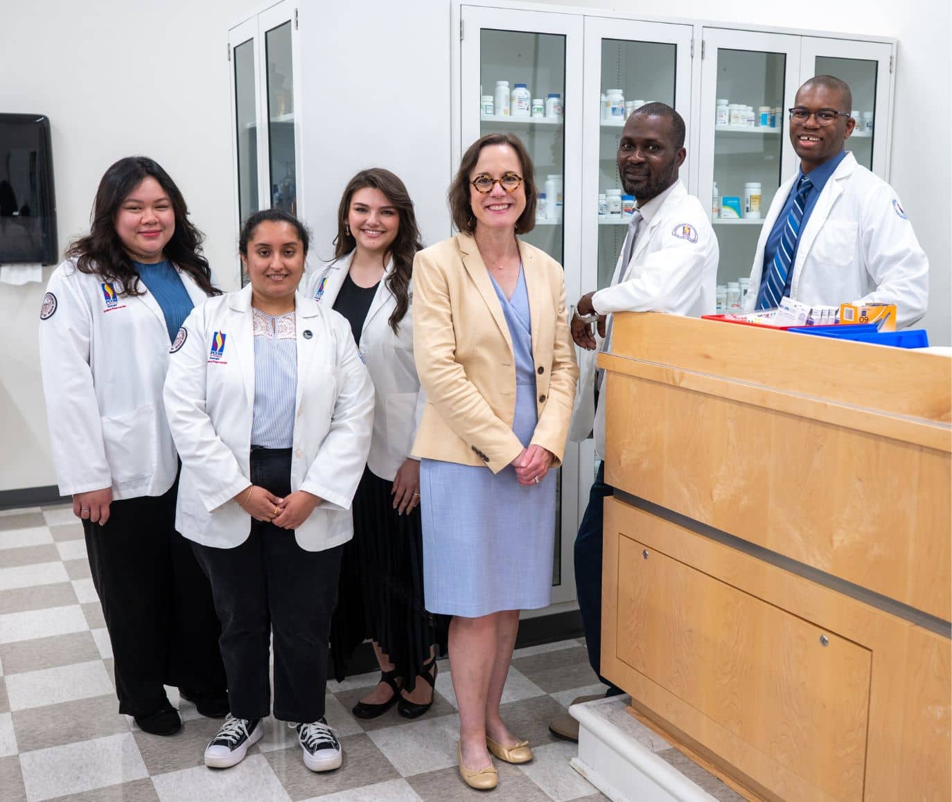 Four pharmacy students in white coats and two faculty members pose together in a PCOM pharmacy lab