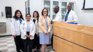 Four pharmacy students in white coats and two faculty members pose together in a PCOM pharmacy lab