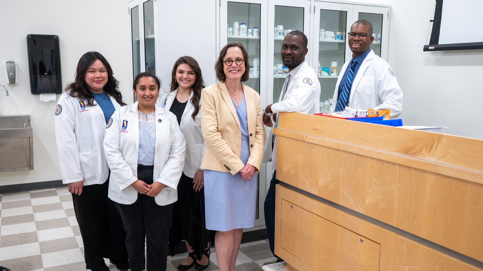 Four pharmacy students in white coats and two faculty members pose together in a PCOM pharmacy lab