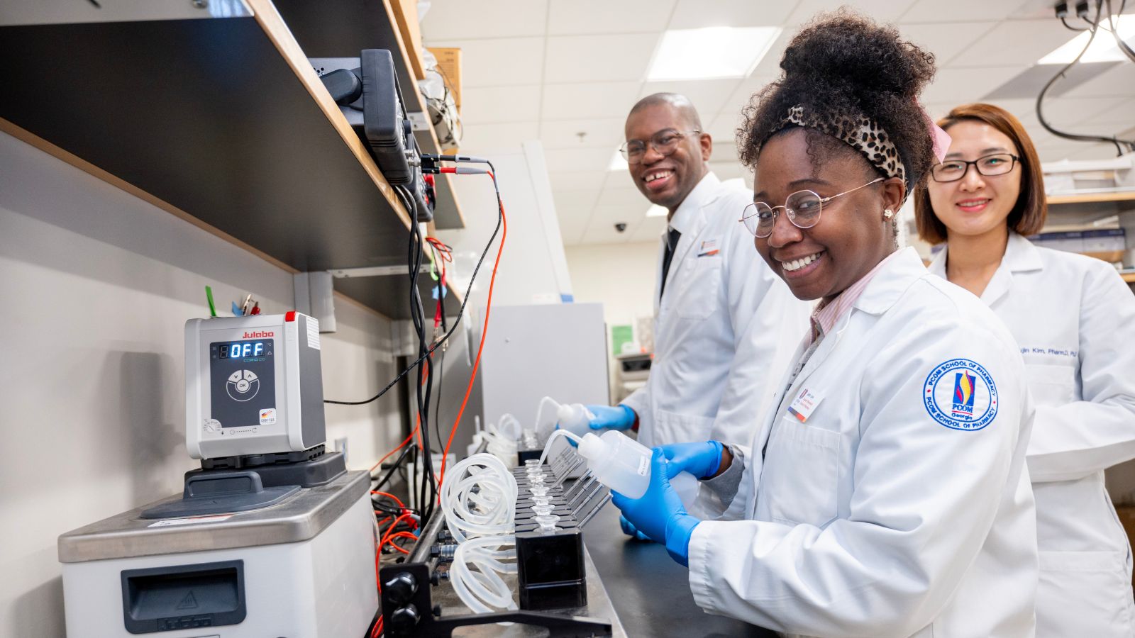 Three PCOM pharmacy students in white coats and blue gloves smile while working with lab equipment at a research bench