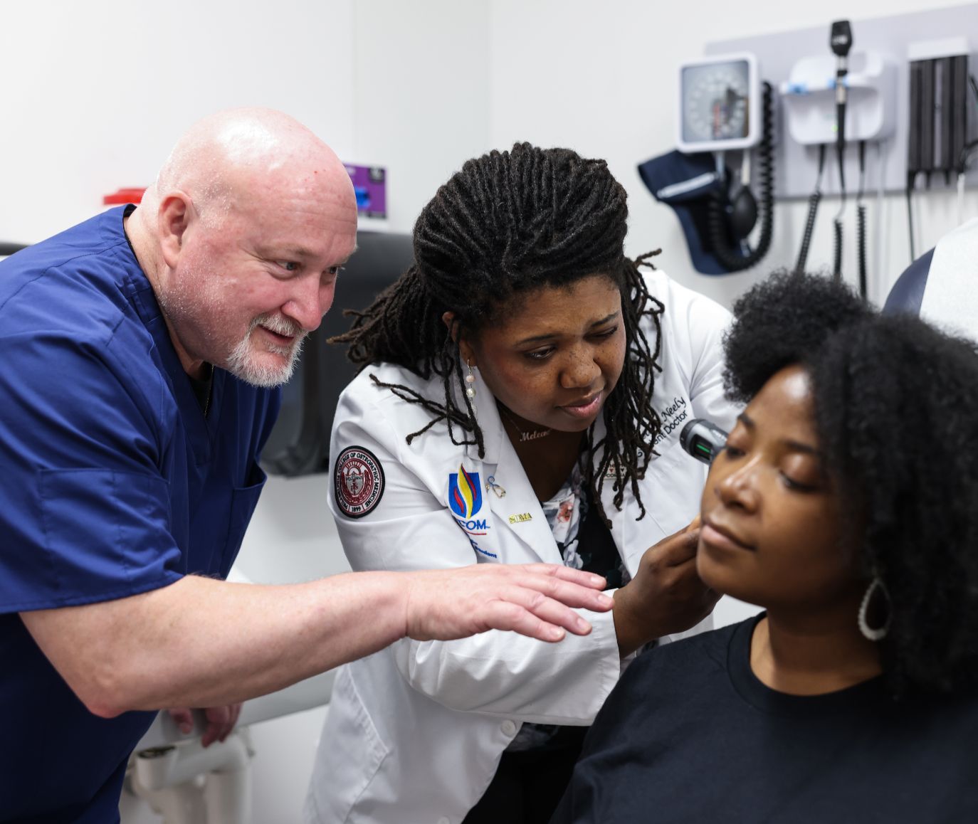 PCOM student doctor examines a patient's ear with an otoscope while an instructor in scrubs observes in a clinical exam room