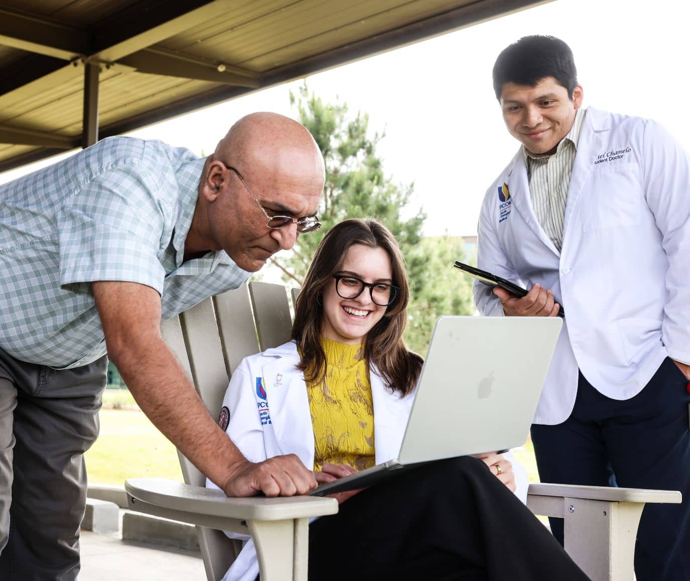 Two PCOM student doctors in white coats and a faculty member gather around a laptop outdoors on campus