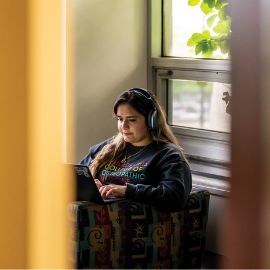 Student wearing headphones works on a laptop while sitting by a window in a campus lounge