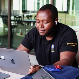Student in a PCOM orientation t-shirt works on a laptop with earbuds in at a campus study area