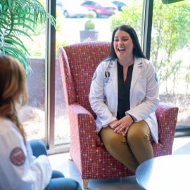 Medical student in a white coat laughing while seated in a lounge chair during a conversation with a colleague