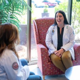 Medical student in a white coat laughing while seated in a lounge chair during a conversation with a colleague