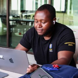Student in a PCOM orientation t-shirt works on a laptop with earbuds in at a campus study area