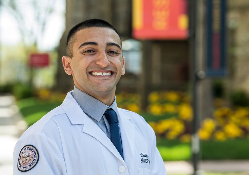 Smiling student physician in a white coat and tie standing outdoors on the PCOM campus with banners and flowers in the background