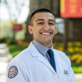 Smiling student physician in a white coat and tie standing outdoors on the PCOM campus with banners and flowers in the background