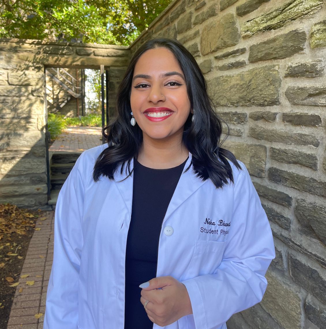 Smiling student physician in a white coat standing outdoors next to a stone wall on the PCOM campus