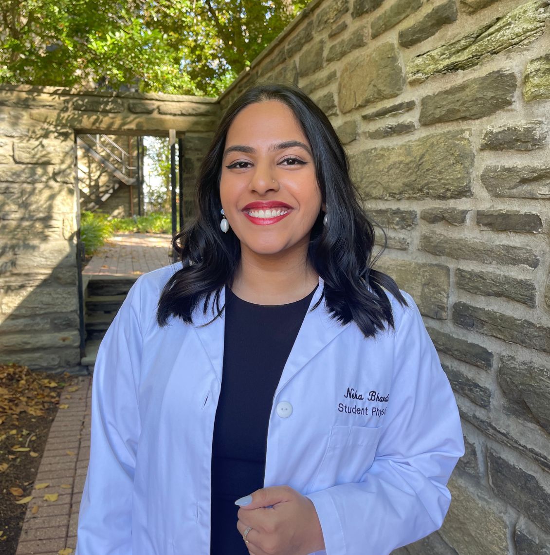 Smiling student physician in a white coat standing outdoors next to a stone wall on the PCOM campus