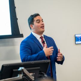 Man in a blue suit gestures while giving a presentation at a podium in a classroom