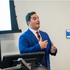 Young man in a blue suit and red tie gestures while presenting at a podium in a classroom setting