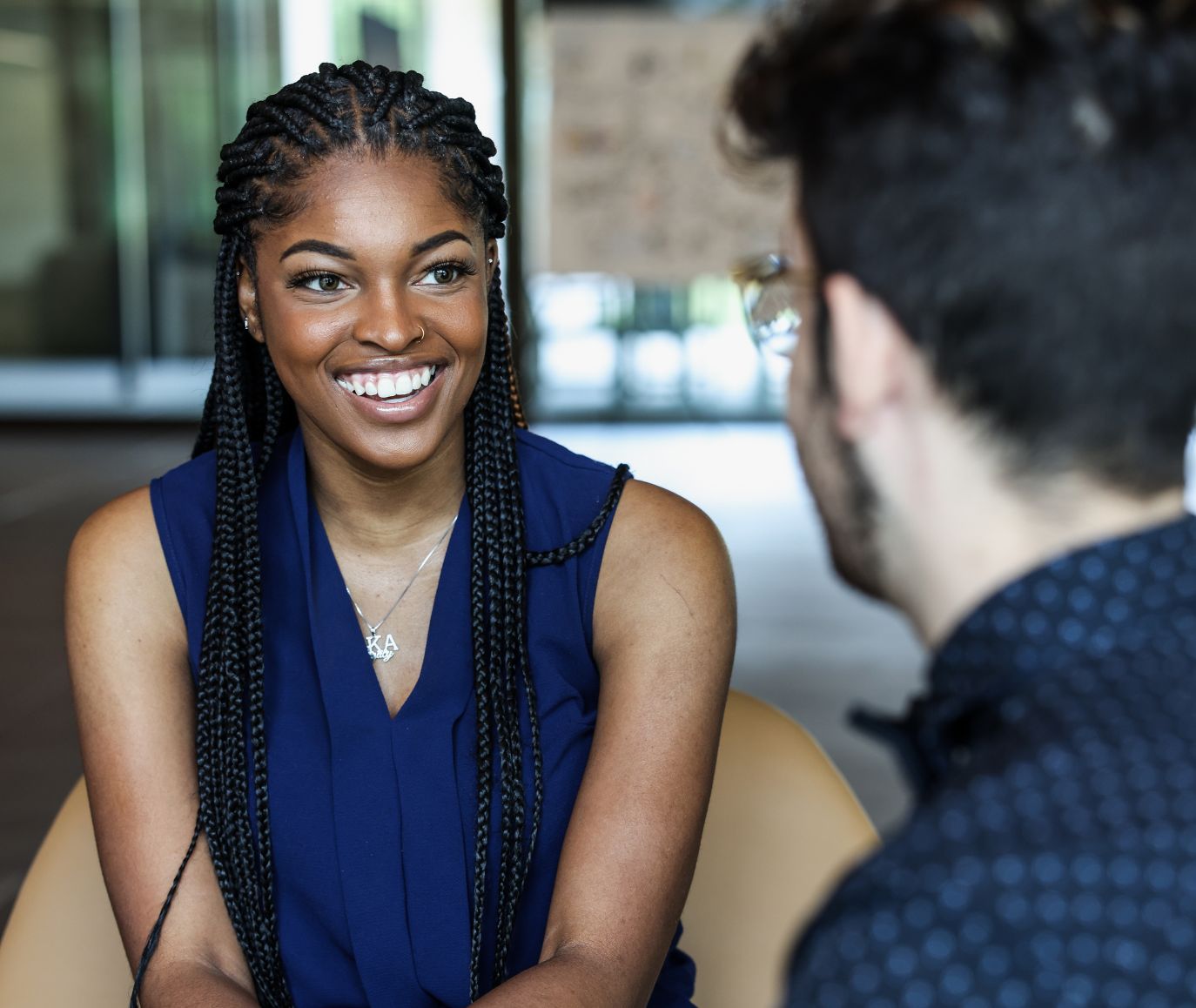 Young woman with braids in a navy blouse smiling during a one-on-one conversation in a modern campus lounge