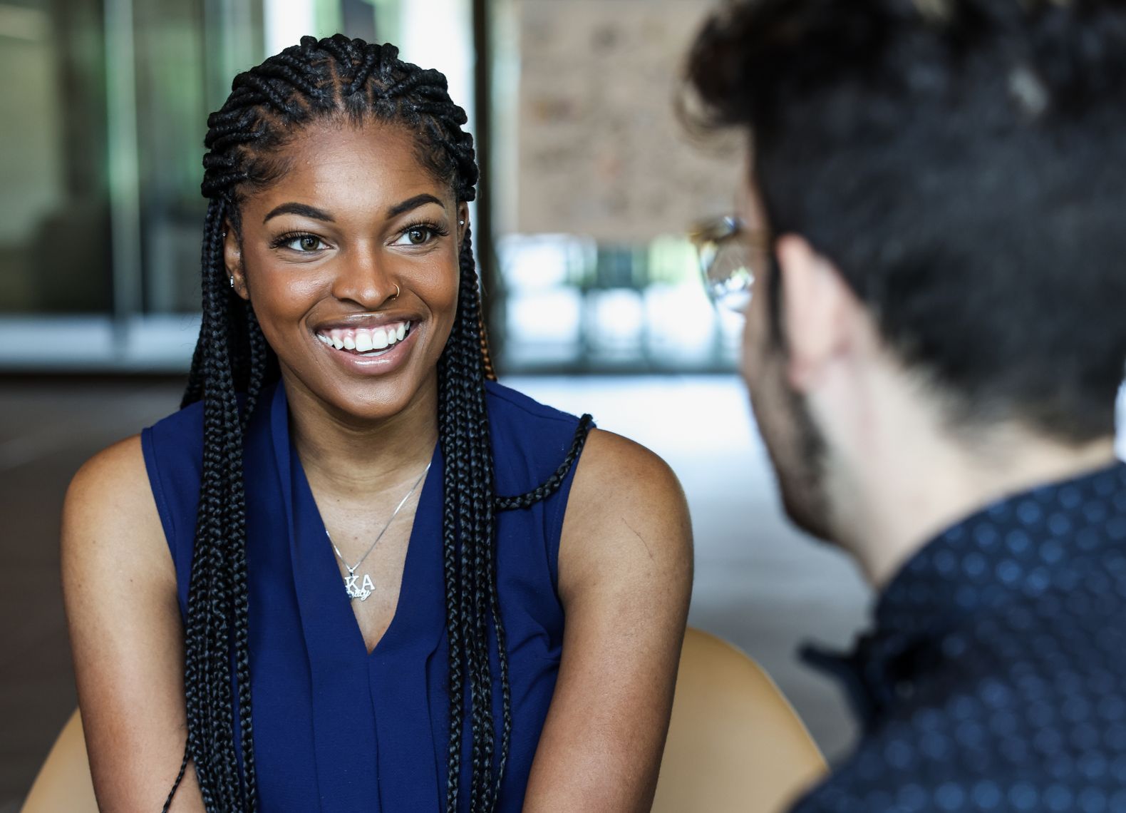 Young woman with braids in a navy blouse smiling during a one-on-one conversation in a modern campus lounge