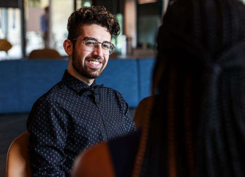 Young man with curly hair and glasses smiling during a conversation in a modern campus lounge