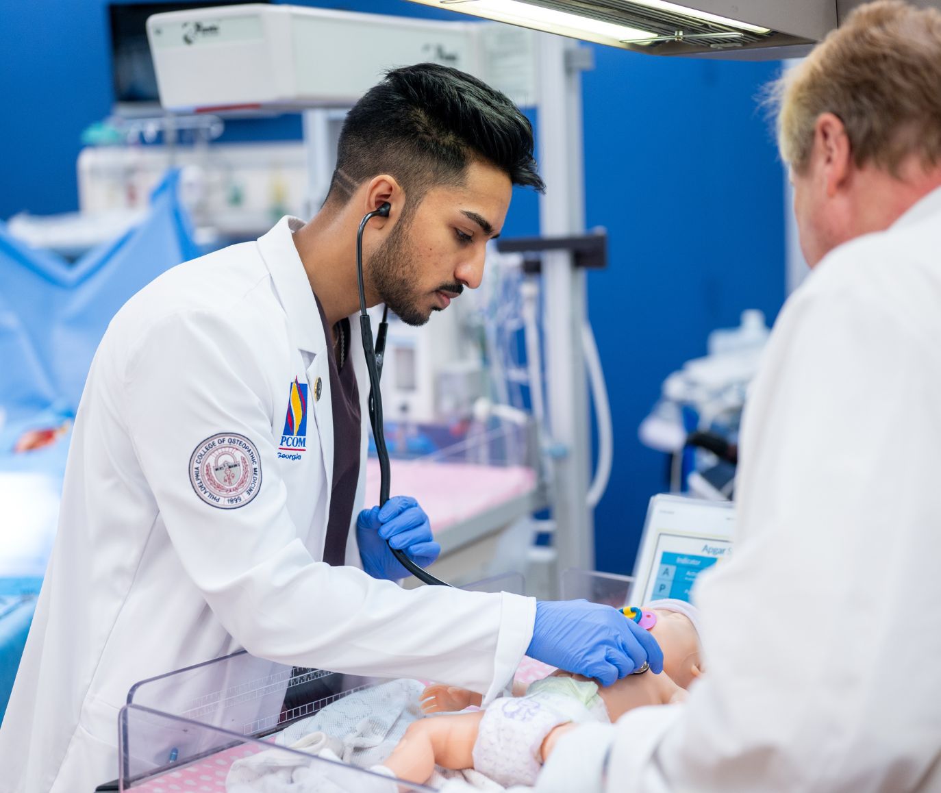 PCOM medical student uses a stethoscope to examine an infant mannequin in a simulation lab while an instructor observes