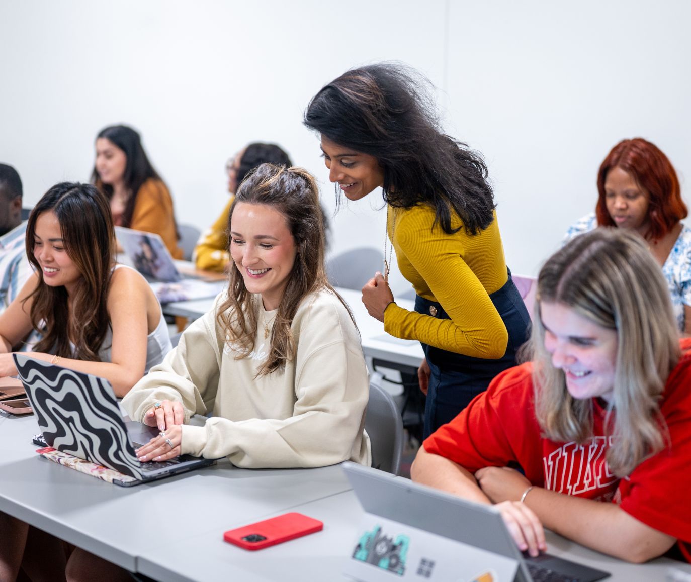 Students smile and work on laptops in a classroom while an instructor leans in to assist