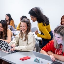 Students laugh and work on laptops in a classroom as an instructor stands nearby offering guidance
