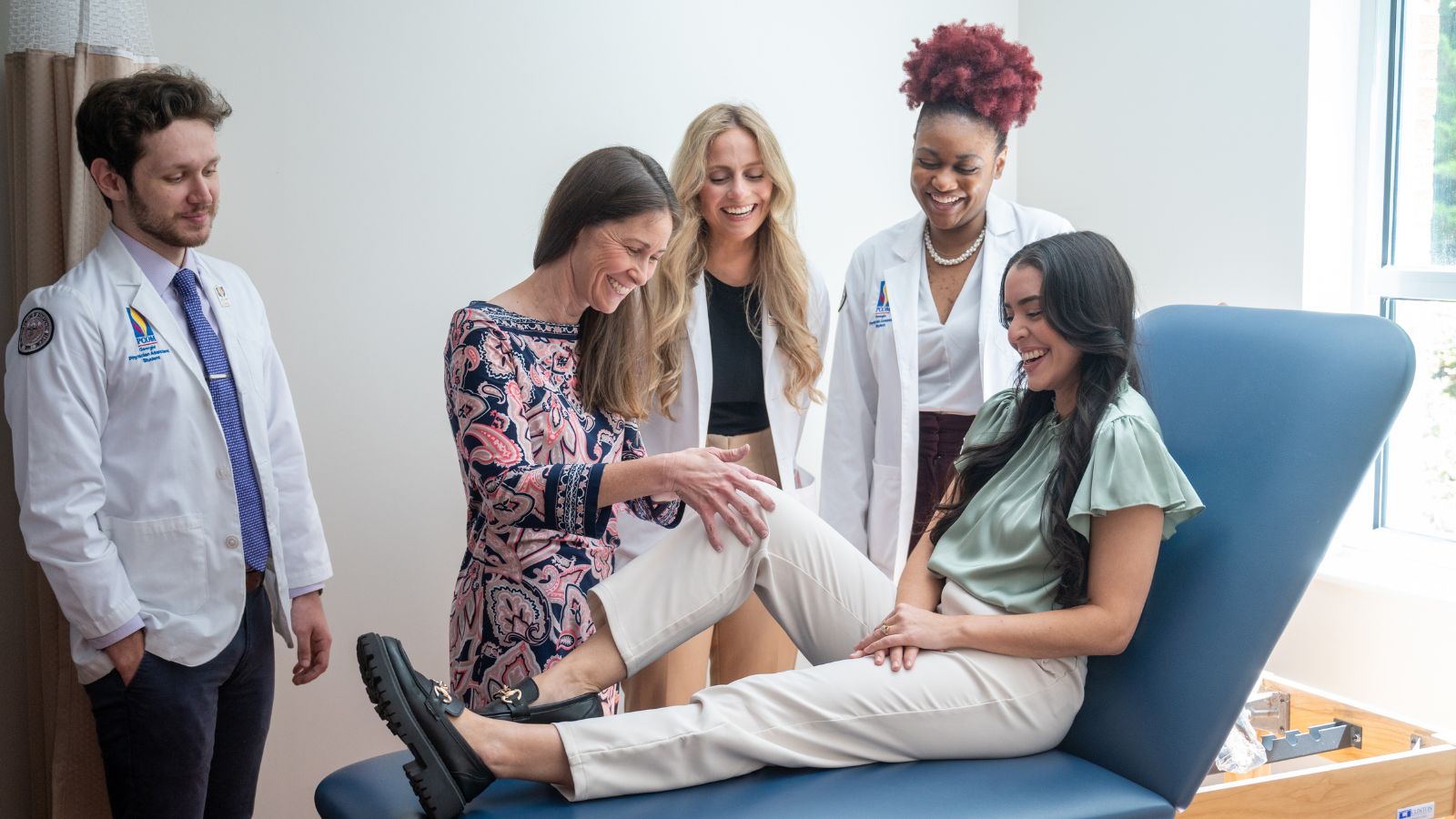 An instructor demonstrates a knee examination on a student seated on an exam table while three PCOM students in white coats observe and smile