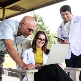 Two students in white coats and a faculty member review a laptop together at an outdoor covered patio