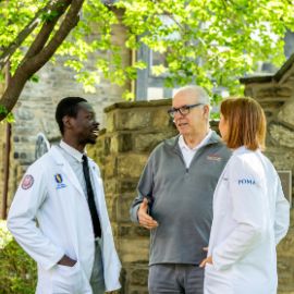 Two students in white coats talk with a faculty member in a PCOM pullover on a tree-lined campus walkway