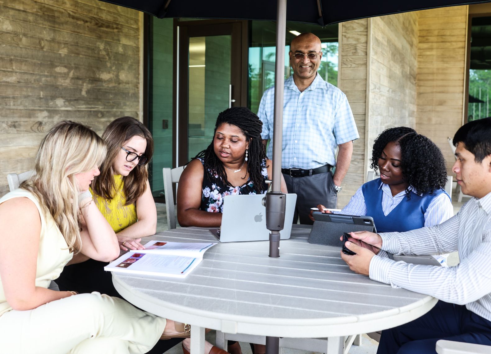 Six students and a faculty member gather around an outdoor patio table with laptops, tablets, and documents