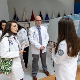 Students and faculty in white coats discuss a research poster at a PCOM academic presentation event