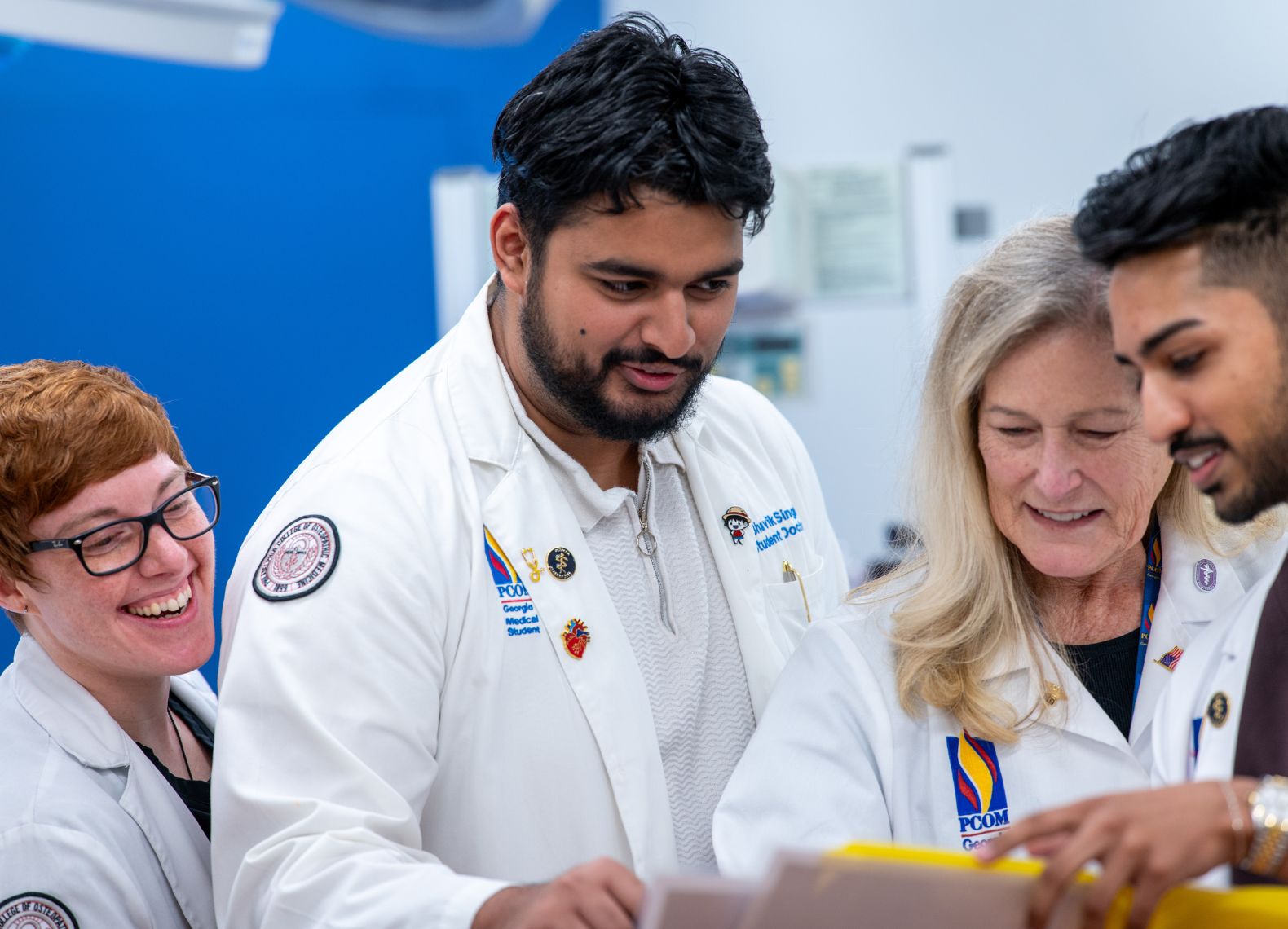 Three PCOM medical students and a faculty member in white coats smile while reviewing a chart together in a simulation lab