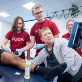 Students in red PCOM shirts observe as an instructor demonstrates a hands-on clinical technique in a training lab