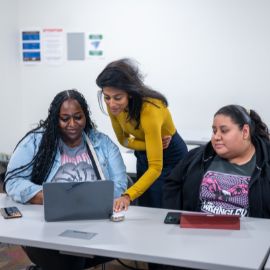 Three students gather around a laptop at a classroom table, with one standing and leaning in to help