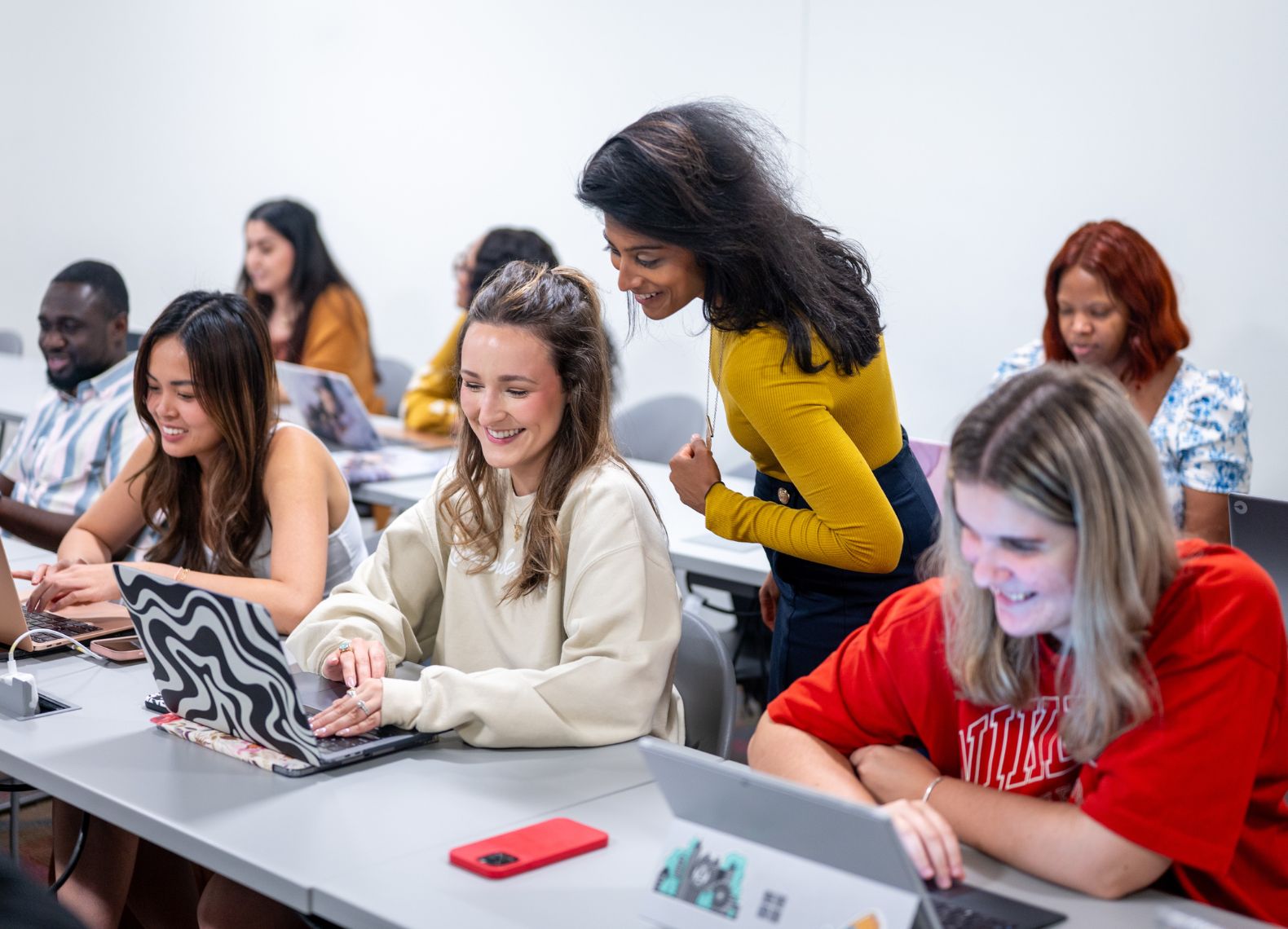 Students smile and work on laptops in a classroom while an instructor leans in to help