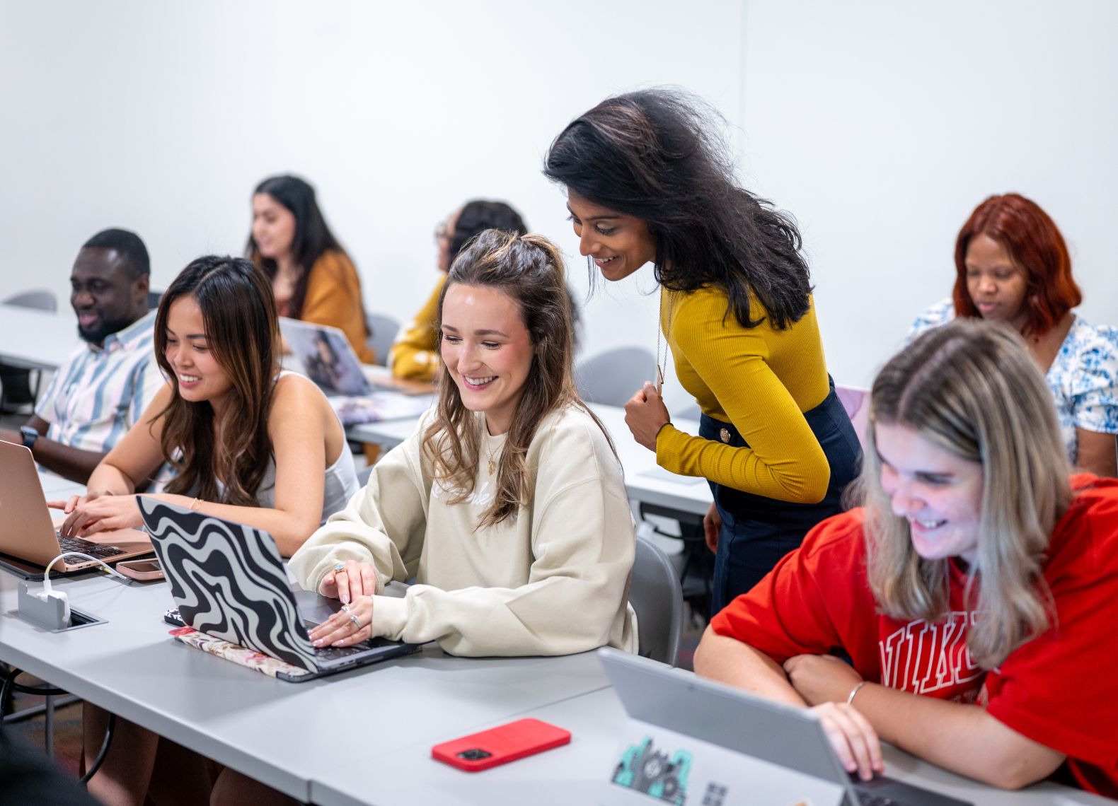 Students smile and work on laptops in a classroom while a peer stands nearby offering guidance