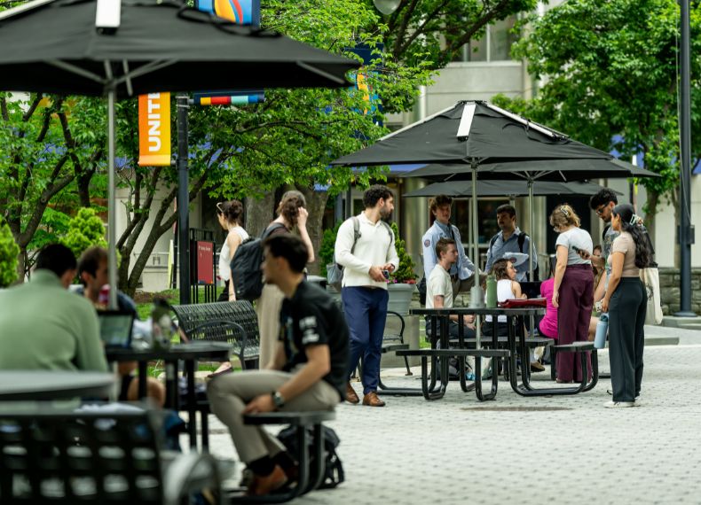 Students gather and socialize at tables with umbrellas in an outdoor campus plaza on a sunny day