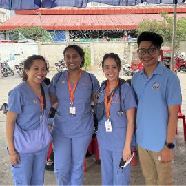 Four students in blue scrubs smile together at an outdoor medical mission site