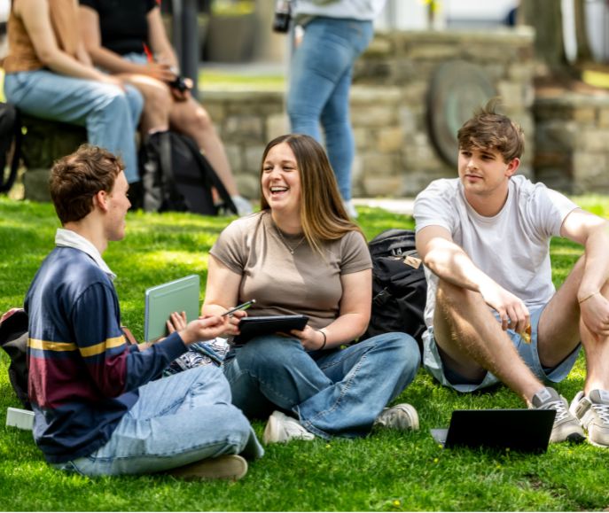 Three students sit on a sunny campus lawn laughing and studying with notebooks and a laptop