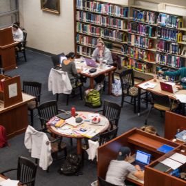 Students study at tables with laptops and books in a campus library with bookshelves in the background
