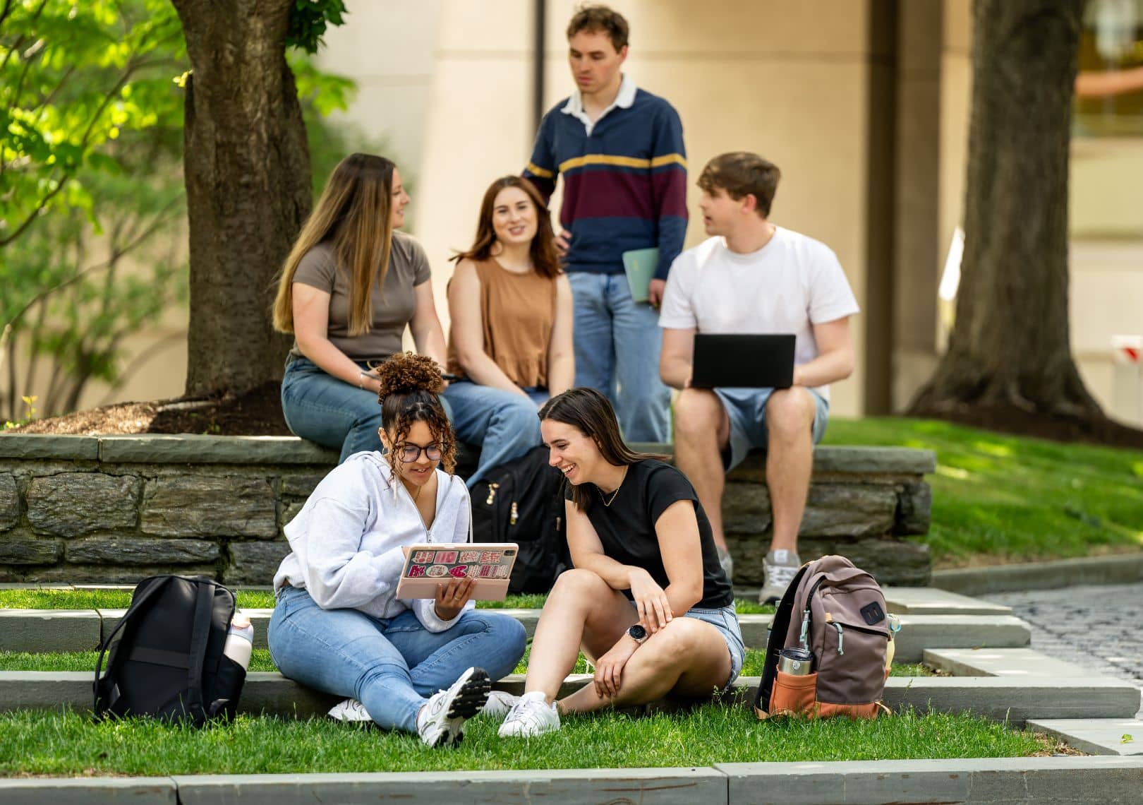 Six students gather on a grassy campus lawn and stone steps, chatting and studying with tablets and laptops