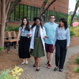 Four students walk together along a tree-lined brick pathway on the PCOM campus, one wearing a white coat